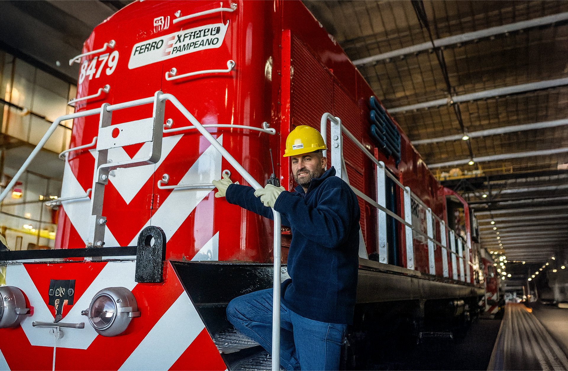 Trabajador ferroviario con casco de seguridad posando junto a una locomotora roja de Ferroexpreso Pampeano dentro de taller.