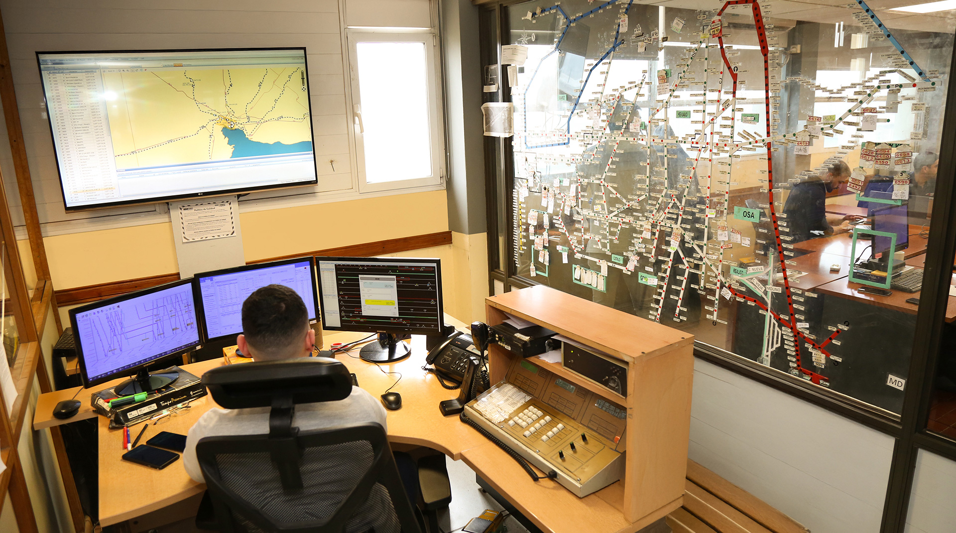 Centro de control ferroviario con operador supervisando pantallas de monitoreo y panel de comunicaciones, frente a un mapa de la red de vías.